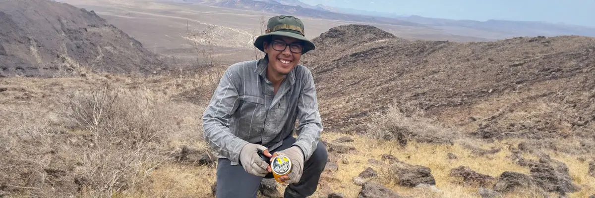 Jose Torres kneeling on a rocky hillside in a desert landscape, wearing a hat and holding a colorful object.